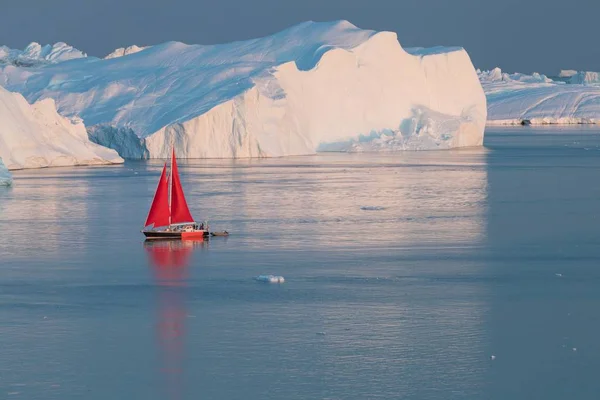 Küçük kırmızı yelkenli polar yaz gece yarısı güneş sezonu sırasında Disko Bay buzul yüzen buzdağları arasında seyir. Ilulissat, Grönland. Antarktika denizinde buzdağı ile sefer gemisi