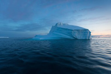 Gün batımında ilginç ve mavi bir gökyüzü altında fotojenik ve karmaşık buzdağı. Doğada küresel ısınmanın etkisi. Antarktika veya Grönland'da derin mavi suda eriyen buzulun kavramsal görüntüsü