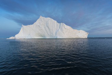 Gün batımında ilginç ve mavi bir gökyüzü altında fotojenik ve karmaşık buzdağı. Doğada küresel ısınmanın etkisi. Antarktika veya Grönland'da derin mavi suda eriyen buzulun kavramsal görüntüsü