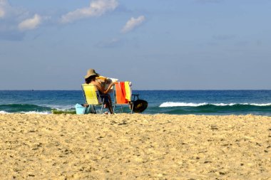 İsrail - Netanya, 14 Ekim 2017: Yetişkin bir kadın Akdeniz kıyısında bir sahil sandalye üzerinde plaj şemsiyesi altında oturur ve bir gazete okuyor