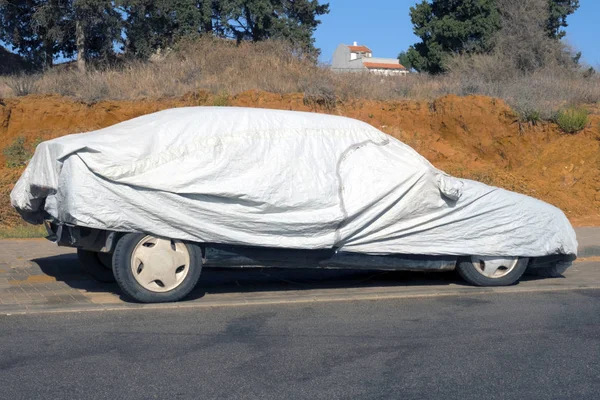 the car covered with a cover stands on the street under the sun