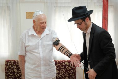 ISRAEL - Netanya, 23 May 2019: an elderly Jewish man puts on a tefillin before a prayer clipart