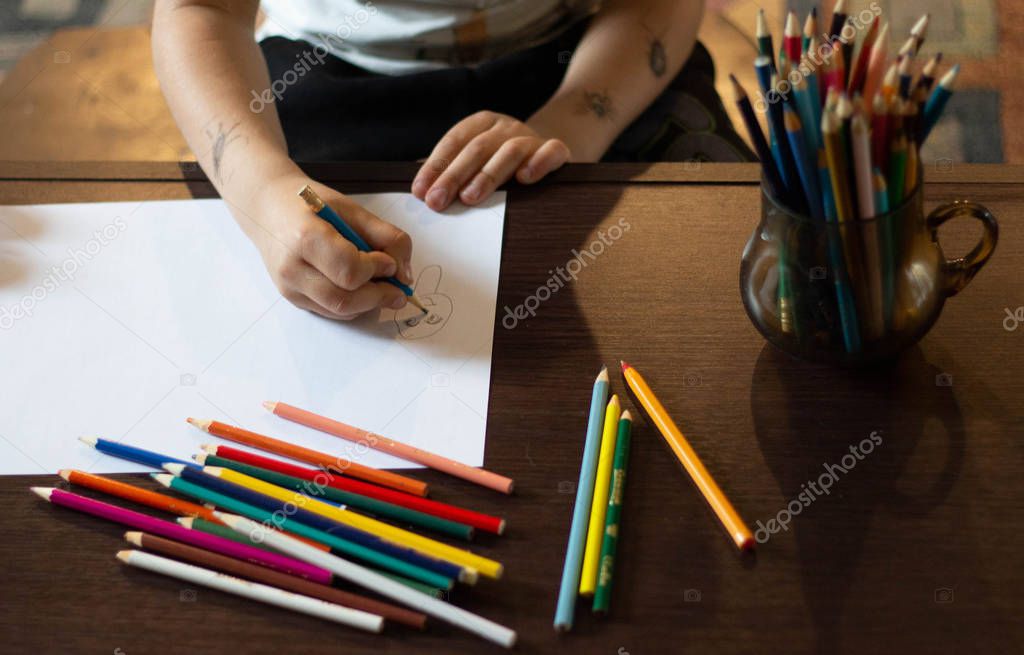 The child's hands are painted with colored pencils on a white sheet of paper on a wooden table.