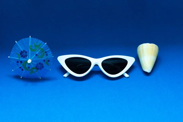 Коктейльный зонтик, белые солнцезащитные очки и панцирь на ярко-голубом фоне. Концепция летнего морского отдыха . — стоковое фото