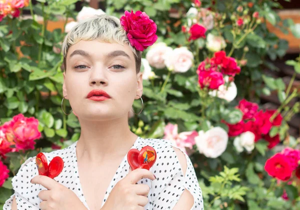 A beautiful young blonde girl in a white blouse and with a rose in her hair expresses emotions in a rose garden on the background of an amazing summer. The girl plays with red castanets.