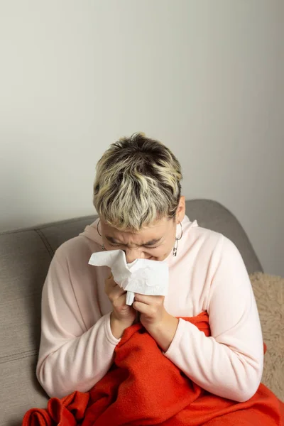 Unhealthy girl sitting on the couch, wrapped in a warm orange blanket, holding a napkin, sneezing and blowing her nose. Girl looks with chronic allergic rhinitis or cold difficult to breathe concept.