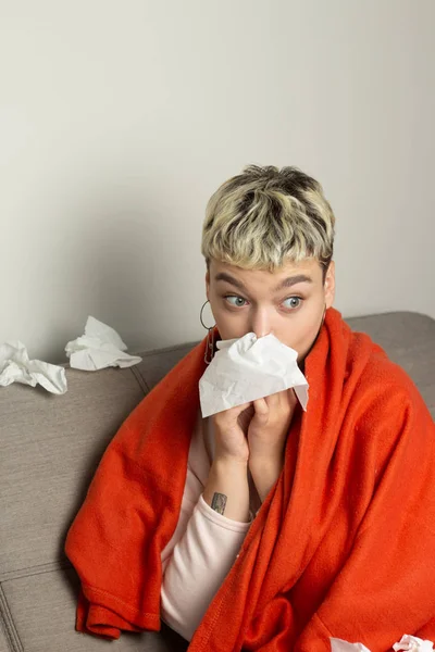 Unhealthy girl sitting on the couch, wrapped in a warm orange blanket, holding a napkin, sneezing and blowing her nose. Girl looks with chronic allergic rhinitis or cold difficult to breathe concept.