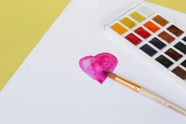 Workspace of the artist with watercolors and brush in the home office. Heart shape drawn with red watercolors on white paper. Valentine\'s Day.