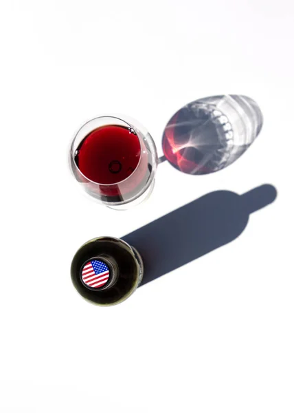 Une bouteille de vin et un verre de vin rouge projettent des ombres dures sur une table blanche. Le drapeau des États-Unis est représenté sur le capuchon de la bouteille. — Photo