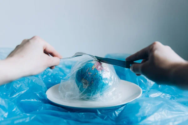 Planet Earth covered in plastic on a plate with a fork and knife eaten up by a human. Plastic pollution and waste concept. Human Impact - Refuse Plastic. Global warming problem.