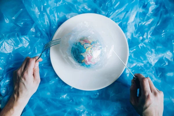 Planet Earth covered in plastic on a plate with a fork and knife eaten up by a human. Plastic pollution and waste concept. Human Impact - Refuse Plastic. Global warming problem.