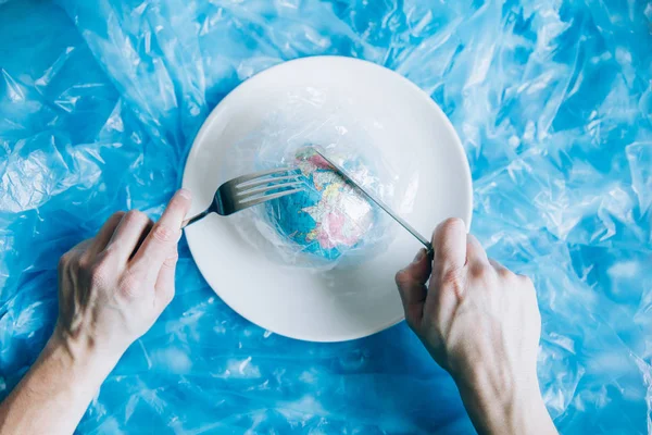 Planet Earth covered in plastic on a plate with a fork and knife eaten up by a human. Plastic pollution and waste concept. Human Impact - Refuse Plastic. Global warming problem.