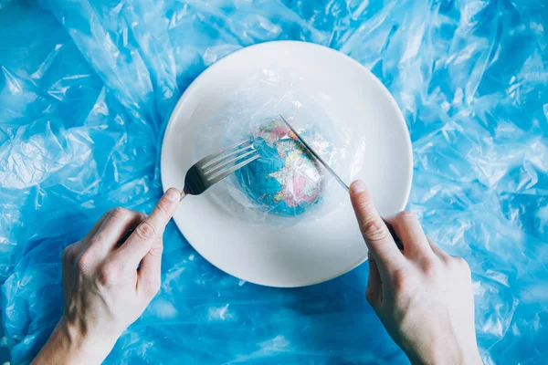 Planet Earth covered in plastic on a plate with a fork and knife eaten up by a human. Plastic pollution and waste concept. Human Impact - Refuse Plastic. Global warming problem.