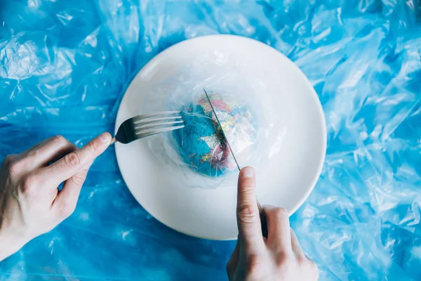 Planet Earth covered in plastic on a plate with a fork and knife eaten up by a human. Plastic pollution and waste concept. Human Impact - Refuse Plastic. Global warming problem.