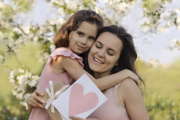 happy mother\'s Day! child daughter gives mom a heart card and a gift. Happy Birthday. People, holidays and family concept-happy daughter and mother against the background of blooming flowers