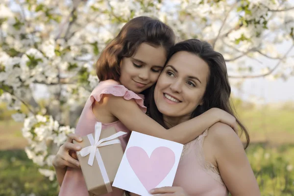 happy mother\'s Day! child daughter gives mom a heart card and a gift. Happy Birthday. People, holidays and family concept-happy daughter and mother against the background of blooming flowers