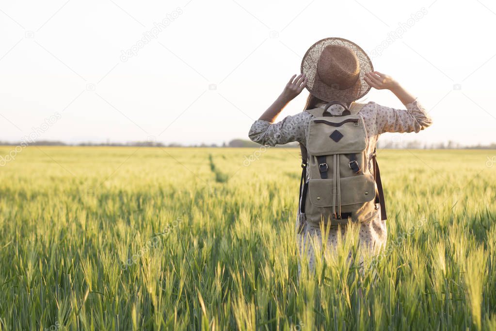 Traveler with a backpack in a field of wheat at sunset. Woman outdoor in nature at the backdrop of the open horizon. The concept of freedom and discovery. Environment and lifestyle