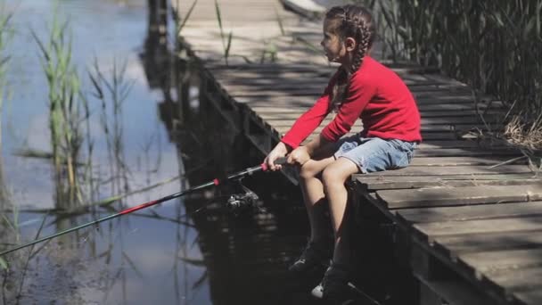 Barn fiske flicka på sjön vid piren. Kastar och drar fiskespö spinning. — Stockvideo