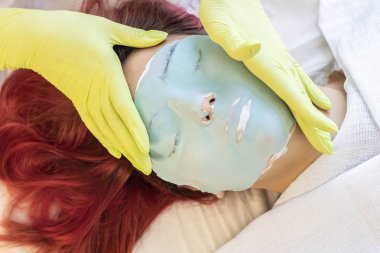 Eller, güzellik uzmanı kızıl saçlı güzel bir kızın yüzüne yüz maskesi taktı. Yüz bakımı konsepti, yaşam tarzı, güzellik hizmetleri, modern moda, blog, notlar
