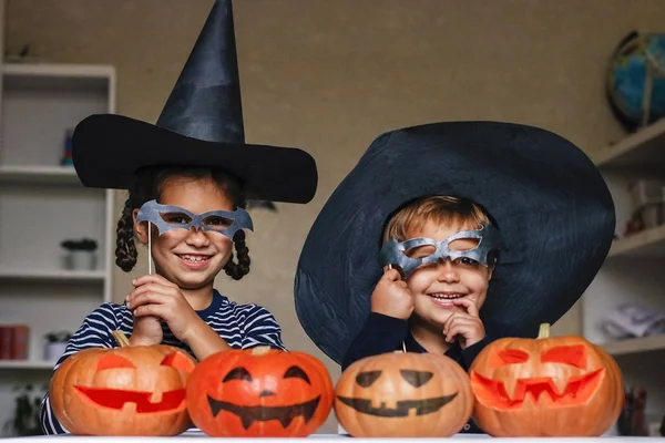 Happy brother and sister celebrate Halloween. Funny children in carnival costumes indoors at the table with pumpkins. Cheerful children play with pumpkins and masks on sticks