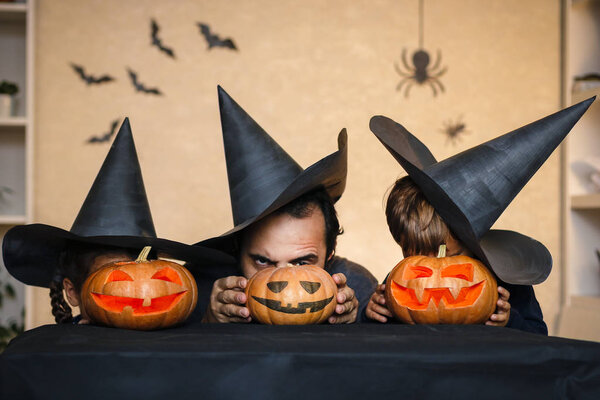 Happy family: father, brother and sister celebrate Halloween. Cheerful children in carnival costumes indoors. Cheerful children and parents play with pumpkins and black witch hats