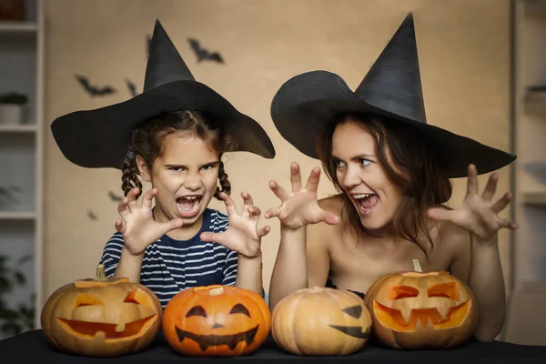 Happy family: mom and daughter celebrate Halloween. Merry people in carnival costumes in the room. Cheerful kids and parents play with pumpkins and black witch hats