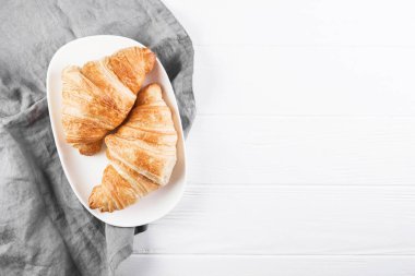 Oval beyaz plaka üzerinde iki kruvasan. Arka planda gri peçete ve beyaz ahşap masa