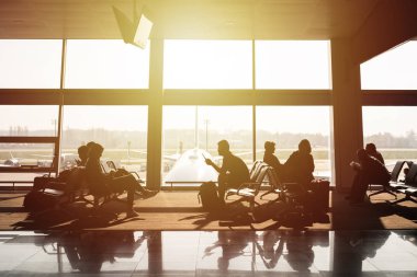 Havaalanı terminali içeride. Yolcular kalkış bekliyor. Arka planda büyük pencere ve arplane.