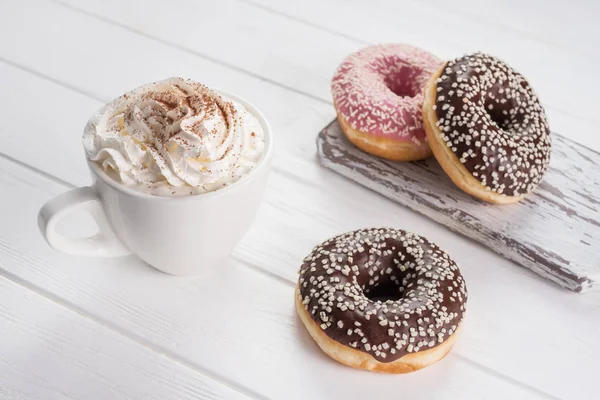 Рожеві та шоколадні пончики з кавою з вершками на білому дерев'яному столі . Ліцензійні Стокові Фото