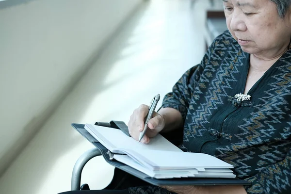 elder woman in wheelchair writing note on notebook near window. elderly mature lifestyle concept