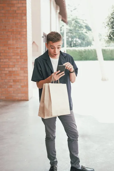 surprised man holding paper shopping bags & mobile smart phone.