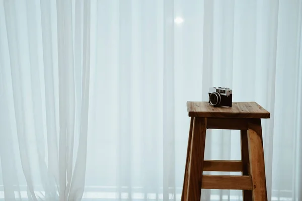 camera on wooden bar stool chair beside see through sheer window curtain