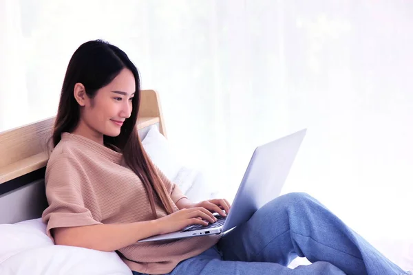 Asian young beautiful  woman sitting on bed using laptop work from home in the morning between virus out break that the worker work at home reduce infection and spreading in many companies