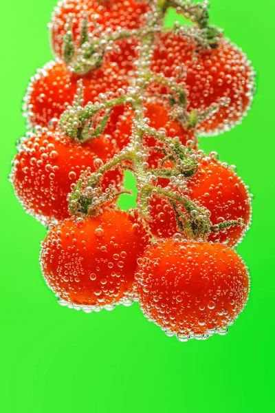 Ripe red cherry tomatoes on branch submerged in water with bubbles. Bright smooth green background.