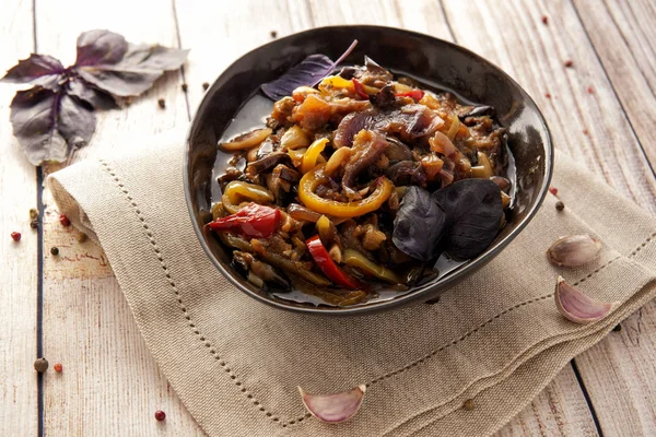 Kalın bir sos içinde haşlanmış aromatik sebzeler. Kafkas mutfağının geleneksel yemeği. Patlıcan, biber ve domates sarımsak ve otlar ile bitkisel yağda. Ketojenik yiyecek. Ahşap bir arka plan ve doğal bir peçete üzerinde bir tabak ta. Yatay görünüm.