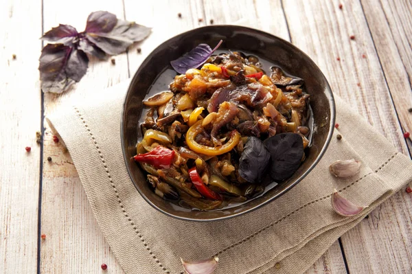 Kalın bir sos içinde haşlanmış aromatik sebzeler. Kafkas mutfağının geleneksel yemeği. Patlıcan, biber ve domates sarımsak ve otlar ile bitkisel yağda. Ketojenik yiyecek. Ahşap bir arka plan ve doğal bir peçete üzerinde bir tabak ta. Yatay görünüm.