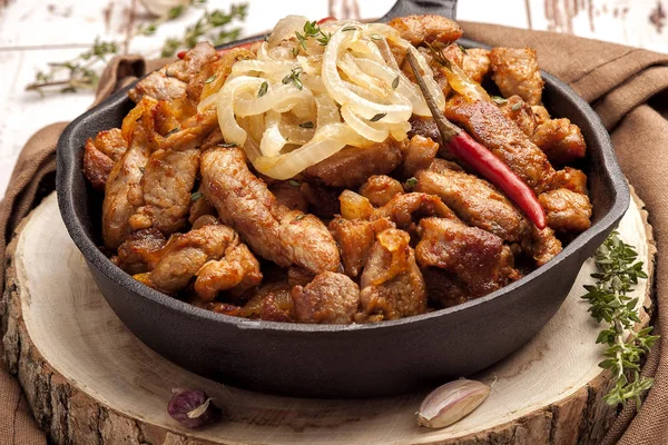 Fried pork with onions and hot pepper in a cast iron pan. Rustic decor. A large portion of meat. Close up and horizontal orientation.