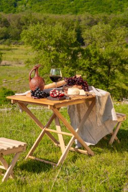 Dağlarda açık hava piknikleri. Kırmızı şarap, peynir, meyve, üzüm ve ekmeğin bulunduğu bir piknik masası yeşil çimlerde bir çayırda duruyor. İzole edilmiş açık hava eğlencesi konsepti.