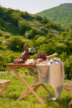Dağlarda açık hava piknikleri. Kırmızı şarap, peynir, meyve, üzüm ve ekmeğin bulunduğu bir piknik masası yeşil çimlerde bir çayırda duruyor. İzole edilmiş açık hava eğlencesi konsepti.
