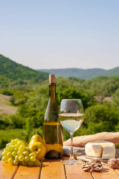 Outdoor picnics in the mountains. A picnic table set with white wine, cheese, fruits, grapes and bread stands in a meadow in green grass. The concept of secluded outdoor recreation