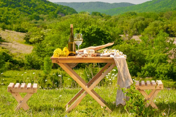 Outdoor picnics in the mountains. A picnic table set with white wine, cheese, fruits, grapes and bread stands in a meadow in green grass. The concept of secluded outdoor recreation