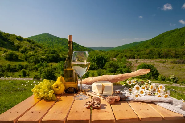 Dağlarda açık hava piknikleri. Beyaz şarap, peynir, meyve, üzüm ve ekmeğin bulunduğu bir piknik masası yeşil çimlerde bir çayırda duruyor. İzole edilmiş açık hava eğlencesi konsepti