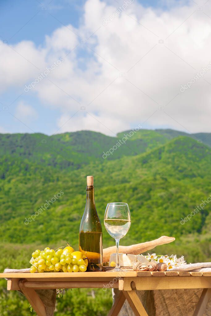 Outdoor picnics in the mountains. A picnic table set with white wine, cheese, fruits, grapes and bread stands in a meadow in green grass. The concept of secluded outdoor recreation