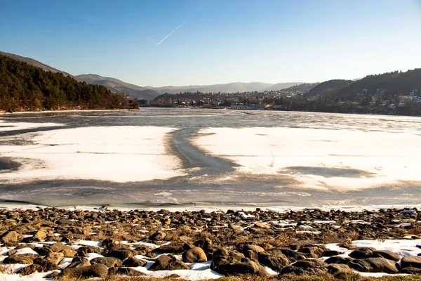 frozen pond with melted thawed patch, stones in the foreground are in the snow, and in the distance are mountains and clear blue sky