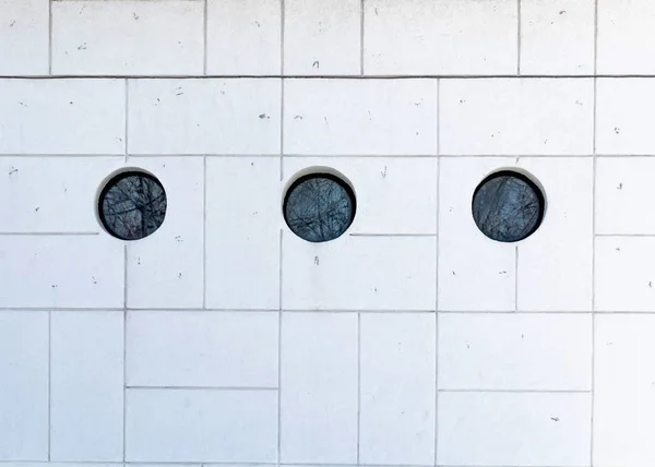 white wall in rectangles and squares and three black round windows with reflections of trees