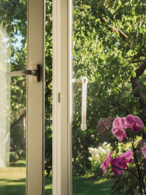 Penceresi biraz açık, pervazında pembe bir orkide var. Eski tarz açık hava termometresi + 22C gösterir. Arka planda yemyeşil bir yaz bahçesi görünüyor..