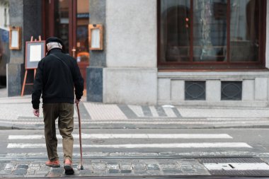 Bastonu olan yaşlı bir erkek, tek başına bir şehrin ortasında bir yaya geçidinde yavaşça yürüyor.