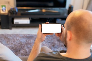 Beyaz beyaz bir erkek arka planda Tv ile moden gün oturma odasında büyük beyaz ekran ile siyah bir akıllı telefon tutar. Telefon izole edilmiş ve yapıştırılan abartmaya hazır.