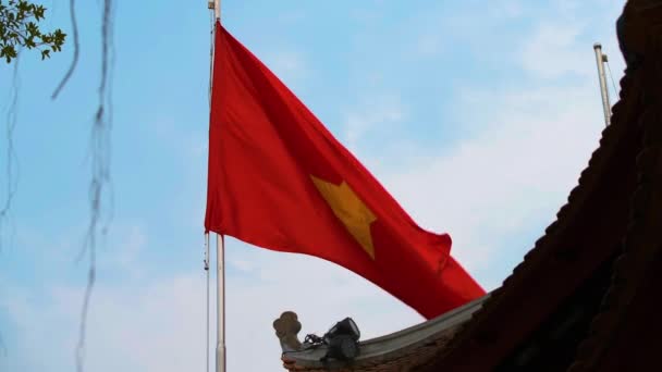 Σημαία Βιετνάμ κυματίζει κατά ένα φωτεινό γαλάζιο ουρανό δίπλα σε ένα ναό στο Ανόι — Αρχείο Βίντεο