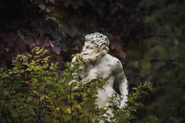 White statue sculpture of Pan the Greek god with a flute in his hand hiding behind foliage looking cheeky